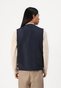 Marineblauwe lichtgewicht vest met een gladde, waterbestendige buitenkant. Beschikt over een mouwloos ontwerp, een hoge kraag en een effen achterkant.