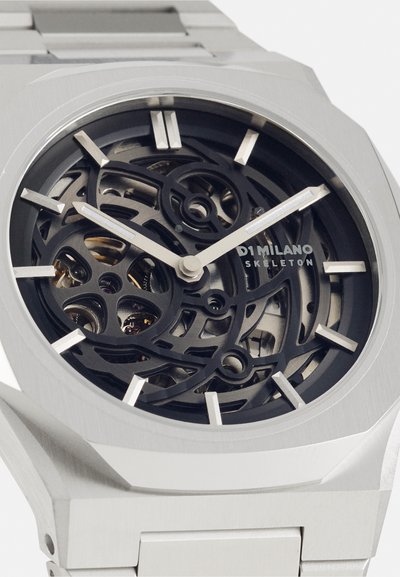 Srebrna ura iz nerjavečega jekla s skeletnim dizajnom, črnim številčnico, izrazitimi kazalci ur in kovinskim zapestjem. Značilne so zapletene notranje mehanike.