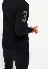 Chemise noire à manches longues avec un col rond, présentant un numéro "3" en relief de couleur grise sur la manche gauche et un ourlet légèrement arrondi.