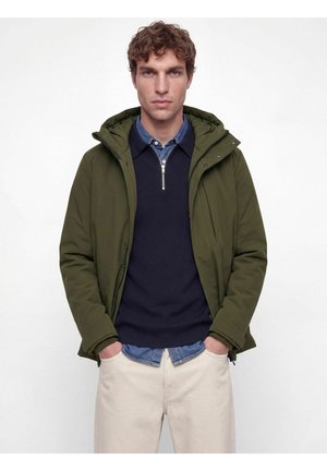 Homme aux cheveux bruns bouclés portant une veste à capuche vert olive sur un pull bleu marine à demi-fermeture éclair et un pantalon beige clair, les mains dans les poches.
