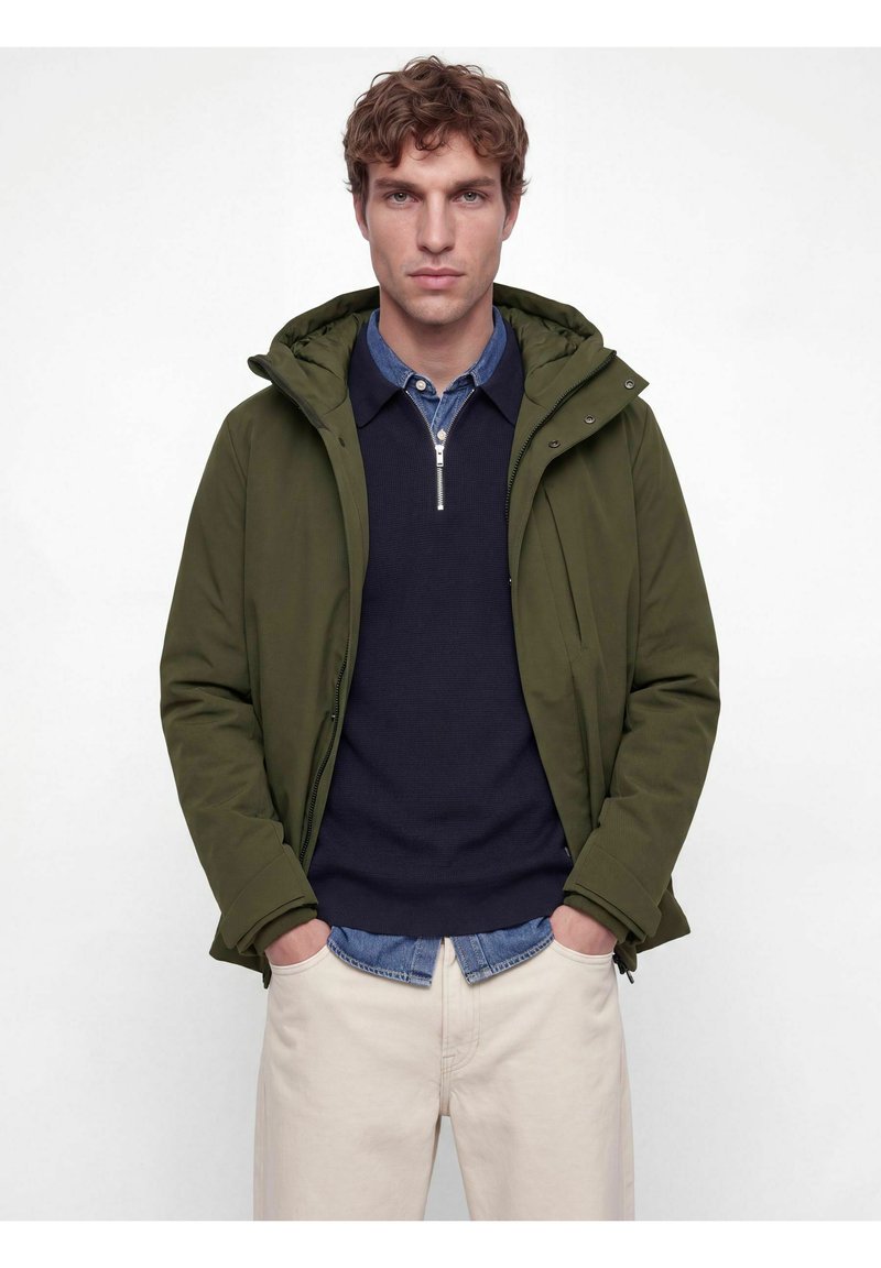 Homme aux cheveux bruns bouclés portant une veste à capuche vert olive sur un pull bleu marine à demi-fermeture éclair et un pantalon beige clair, les mains dans les poches.