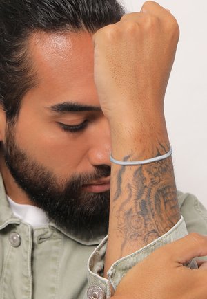 Light blue smooth elastic bracelet worn on a bare wrist, showcasing a detailed tattoo on the forearm. Background is plain.