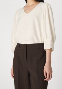 Vrouw in een crèmekleurige V-hals blouse met geborduurde manchetten en donkere, op maat gemaakte trousers, staande tegen een effen achtergrond.