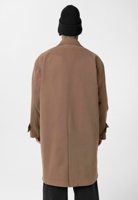 Persoon gekleed in een lange bruine jas en een zwarte gebreide muts, gezien van achteren tegen een effen witte achtergrond.