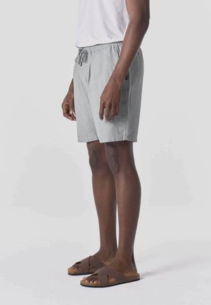 Man staat en draagt lichtgrijze shorts met trekkoord, een wit T-shirt en bruine sandalen met kruisbandjes tegen een egale lichte achtergrond.