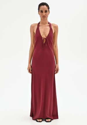Donna dai capelli scuri indossa un abito senza maniche color bordeaux, lungo fino a terra, con scollo all’americana e dettaglio annodato sul davanti, in piedi contro uno sfondo neutro.