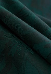 Dunkelgrüner Stoff mit einem strukturierten Blumenmuster. Das Material wirkt gewebt mit subtilen variations in Farbton und Tiefe und zeigt filigrane Details.
