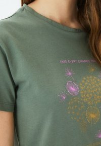 Camiseta de algodón color verde olivo con mangas cortas que presenta un diseño impreso de flores y texto en rosa y dorado. Cuello redondo, textura suave.