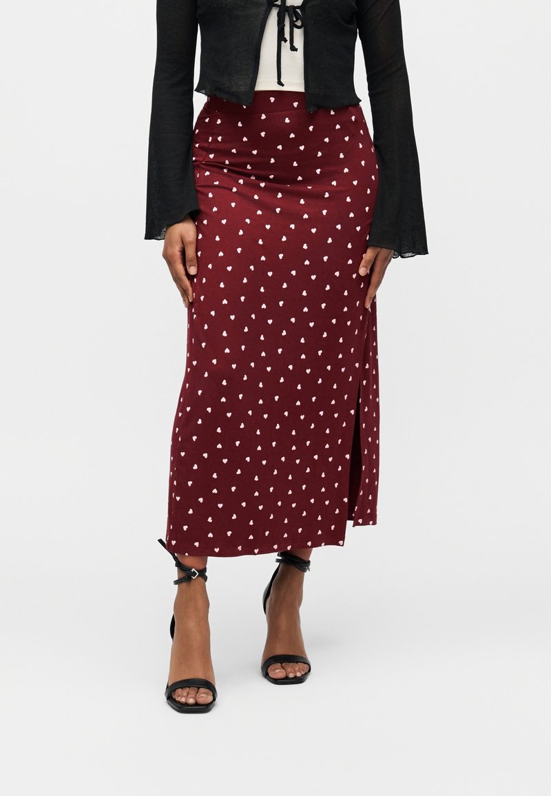 Femme portant une jupe maxi bordeaux avec des motifs de cœurs blancs, un top court noir et des sandales à talons hauts avec bride à la cheville noires.