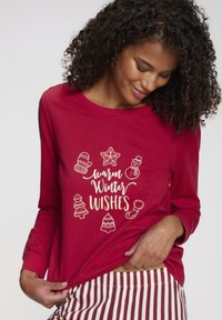 Frau mit lockigem Haar, die ein rotes Langarmshirt mit dem Aufdruck "warme Winterwünsche" und Weihnachtsmotiven trägt und den Saum des Shirts hält.