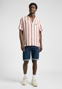 Kurzärmliges, rosa-weiß gestreiftes Hemd mit einer Tasche, kombiniert mit dunkelblauen Jeansshorts und beigen Sneakers. Einfarbiger Hintergrund.