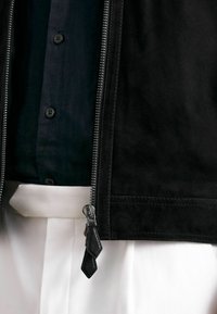 Melna zamšā jaka ar sudraba rāvējslēdzēju, šuvju detaļām un teksturētu rāvējslēdzēja vilkšanas sloksni, apvienota ar tumšu krekla un baltām biksēm.