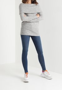 Maglione grigio con spalle scoperte e texture a coste, abbinato a jeans skinny blu. La modella indossa sneakers bianche con accenti arancioni e rosa.