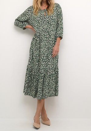Vrouw in een groene midi-jurk met witte bloemenprint, driekwartmouwen, kleine sleutelgatdetail op de borst en beige puntige hakken, staand met één hand op de heup.