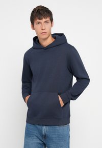 Granatowa bluza z kapturem z mieszanki bawełny, z kieszenią z przodu, ściągaczami na rękawach i luźnym krojem. Noszona z jasnoniebieskimi dżinsami.