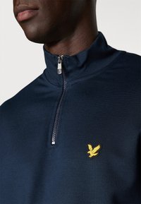 Mėlynos spalvos pusė-užtrauktuku sportinis džemperis su geltonu siuvineštu erelio logotipu ant krūtinės ir sklandžiu užtrauktuku.