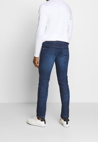 Jeans de mezclilla azul oscuro con un corte ajustado, diseño de cinco bolsillos, costuras sutiles y detalles en contraste, combinados con zapatillas blancas.