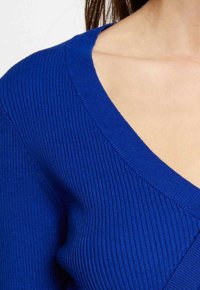 Gros plan sur une personne portant un pull bleu à côtes avec un col en V, montrant le décolleté et les épaules.