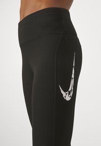 Svarta leggings med en slät textur och en åtsittande design. Har en stor, vit Nike-logga med ett dekorativt mönster på låret.