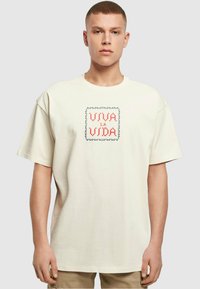 Kremfarget t-skjorte med korte ermer og et firkantet grafisk motiv med "VIVA LA VIDA" i rødt og blått, omgitt av dekorativ søm.