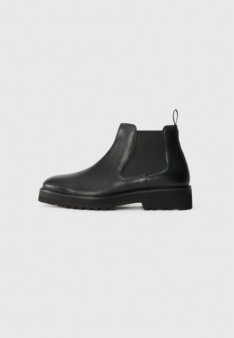 Bota chelsea de cuero negro con panel lateral elástico, puntera redondeada, suela texturizada y pequeña lengüeta en el talón.