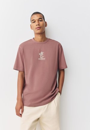 Rust-colored cotton t-shirt with short sleeves, crew neck, and central white graphic text and floral design. Smooth texture, relaxed fit.
