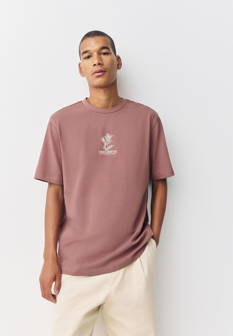 Rust-colored cotton t-shirt with short sleeves, crew neck, and central white graphic text and floral design. Smooth texture, relaxed fit.