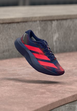 Baskets athlétiques bleues et rouges avec trois bandes rouges, motif floral et lacets noirs flottant au-dessus d'une surface en pierre.