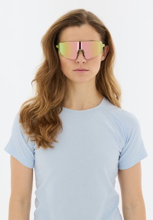 Vrouw met lang golvend haar die neongele en roze reflecterende sportzonnebril draagt en een lichtblauw shirt met korte mouwen, kijkt recht vooruit.