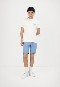 Camiseta de algodón blanca con logo impreso, combinada con pantalones cortos azules claros y zapatillas blancas. El modelo está de pie frente a un fondo liso.