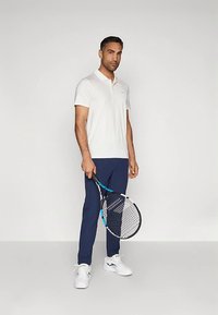 Muž stojící s tenisovou raketou, oblečený v bílém polokošili, tmavě modrých kalhotách a bílých sportovních botách na jednobarevném pozadí.