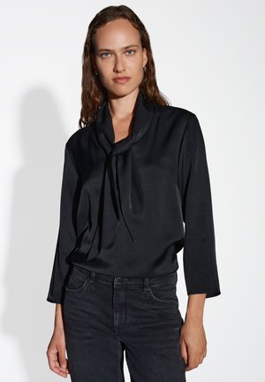 Schwarze Bluse aus glattem Stoff, mit einer gebundenen Schal-Kragen und Dreiviertelärmeln, kombiniert mit dunklen Jeans.
