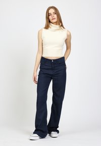 Cremefarbener, gerippter ärmelloser Rollkragenpullover kombiniert mit dunkelblauen Jeans mit weitem Bein. Die Jeans verfügen über Vordertaschen und einen Knopfverschluss. Weiße Sneakers runden den Look ab.