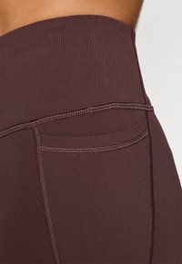 Υψηλής μέσης, σκούρες καφέ κολάν που διαθέτουν έναν λείο, ελαστικό ύφασμα με επίπεδες ραφές και μια μικρή τσέπη στο πίσω μέρος.