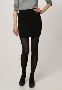 Mini falda negra texturizada combinada con medias opacas negras y tacones de punta con acentos de tachuelas. Parte superior gris de manga larga visible.