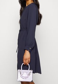 Femme portant une robe bleu marine à manches longues tenant un petit sac à main violet clair avec une chaîne et un motif texturé.