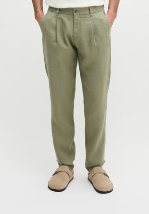 Mann trägt olivgrüne plissierte Hose und beigen Slip-On-Schuh, steht mit Händen in den Taschen vor einem einfarbigen Hintergrund.