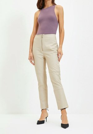 Femme portant un haut sans manches violet, un pantalon beige taille haute avec fermeture éclair à l'avant, et des escarpins noirs à bout pointu, se tenant devant un fond blanc.