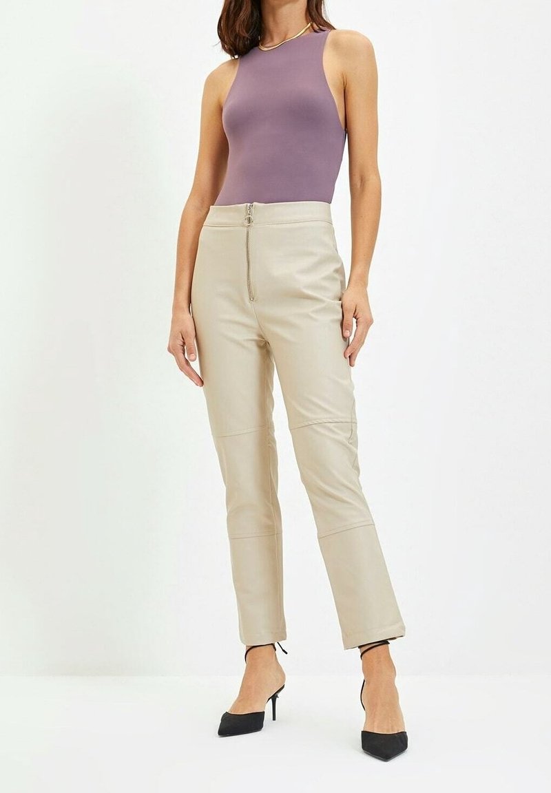Vrouw in een mouwloze paarse top, beige hoge taille broek met rits aan de voorkant, en zwarte puntige hakken, staande voor een witte achtergrond.