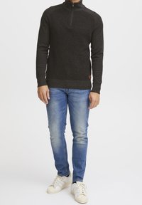 Pull à fermeture éclair gris foncé avec tissu texturé, poignets et ourlet côtelés, associé à un jean bleu clair et des baskets blanches.