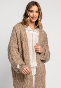Gruba beżowa dziergana kardigan z wzorem w warkocze, z opuszczonymi ramionami i metką na lewym rękawie. Miękka, teksturowana tkanina.