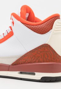 Le sneakers presentano pelle bianca, dettagli arancioni, sezioni perforate, una suola intermedia grigia e un pannello rosso testurizzato. Suola in gomma nera visibile.