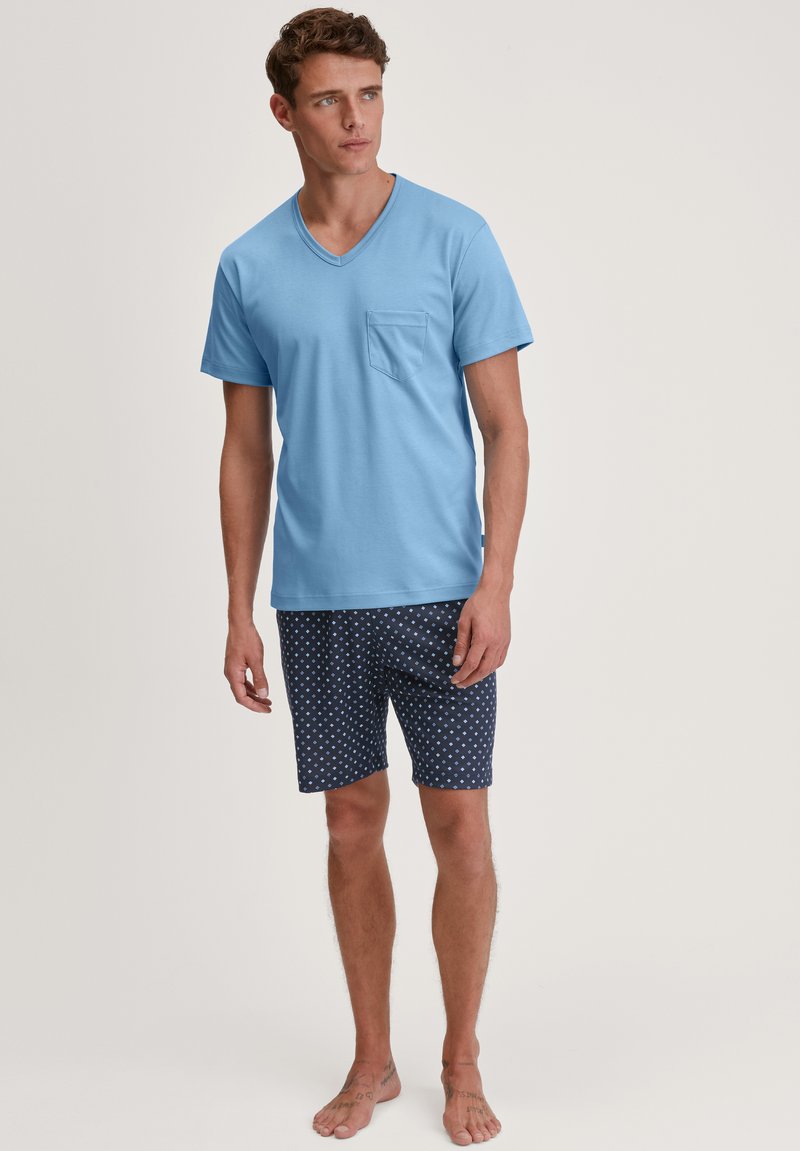 Lichtblauw katoenen V-hals T-shirt met een zak, gecombineerd met marineblauwe shorts met witte stippen, blootsvoets op een neutrale achtergrond.