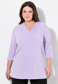Donna con capelli castani fino alle spalle, che indossa orecchini a cerchio dorati e una maglietta viola chiaro a V con maniche a tre quarti.