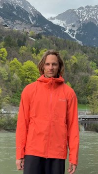 Homme en veste orange vif se tient près d'une rivière avec des arbres verts et des montagnes enneigées en arrière-plan.