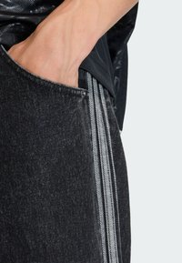 Čierne denimové nohavice so štrukturálnym textúrovaním, obsahujúce bočné vrecko a sivé vertikálne pruhy pozdĺž švov.