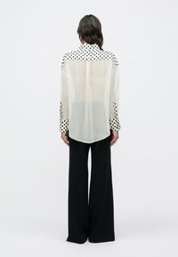 Bluză albă transparentă cu buline negre, manșete cu model contrastant și tiv curbat, asortată cu pantaloni negri largi. Vedere din spate.