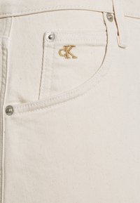 Beige farkkutakki, jossa on teksturoitu pinta, kullanvärisellä brodeeratulla logolla ja hopeisilla osilla. Suunnittelussa on klassinen taskuasettelu.