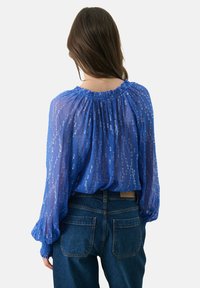 Blaues transparentes Bluse mit gerafftem Ausschnitt und langen, luftigen Ärmeln, die ein strukturiertes Muster aufweist und mit hochgeschnittenen Denim-Jeans kombiniert ist.