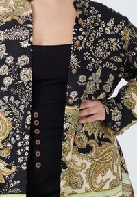 Zwart en crèmekleurig shirt met een patroontje van bloemdecoraties, voorzien van bronzen knopen en een lichtgroene accent aan de zoom, gemaakt van een gladde stof.